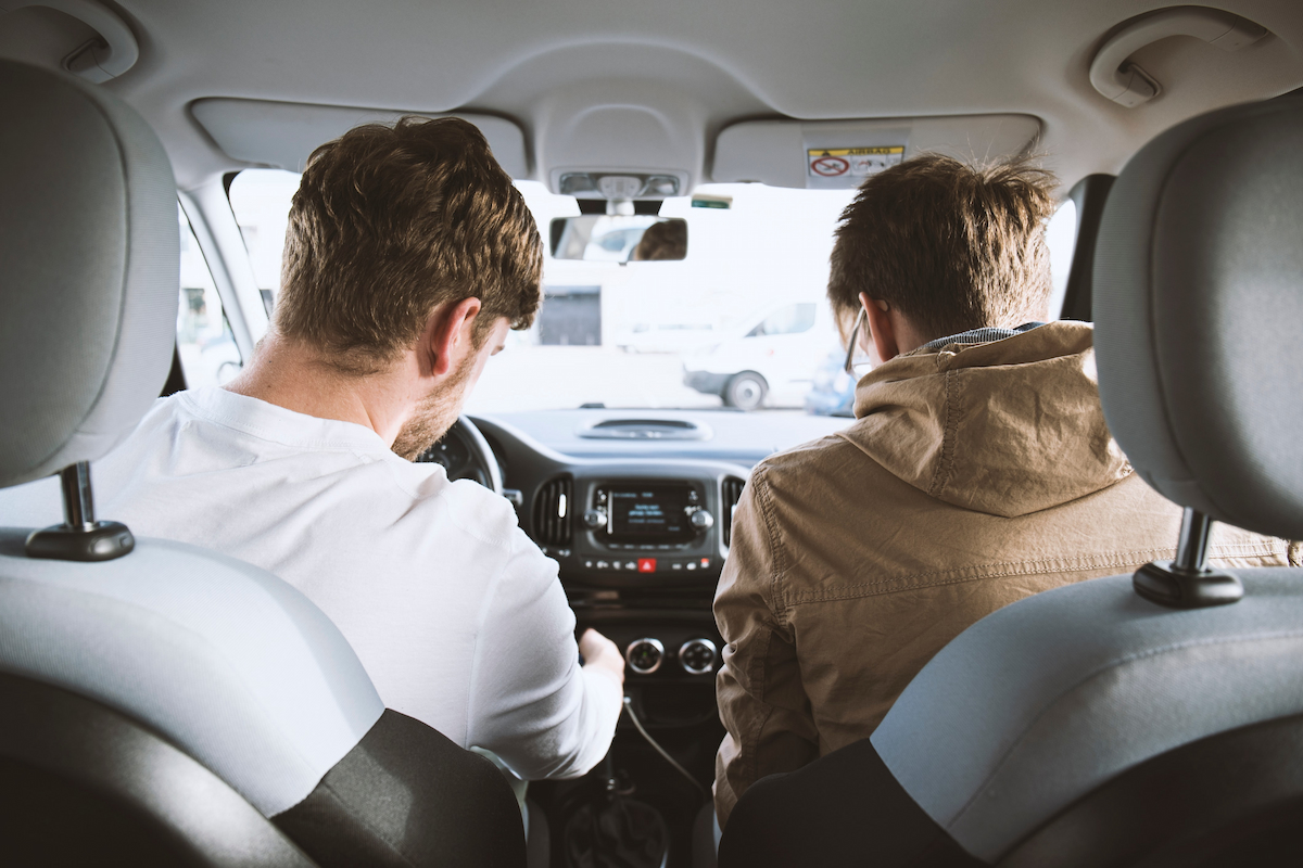 A man wearing a white t-shirt and another man wearing a brown jacket are sitting inside a vehicle looking at the dashboard button.