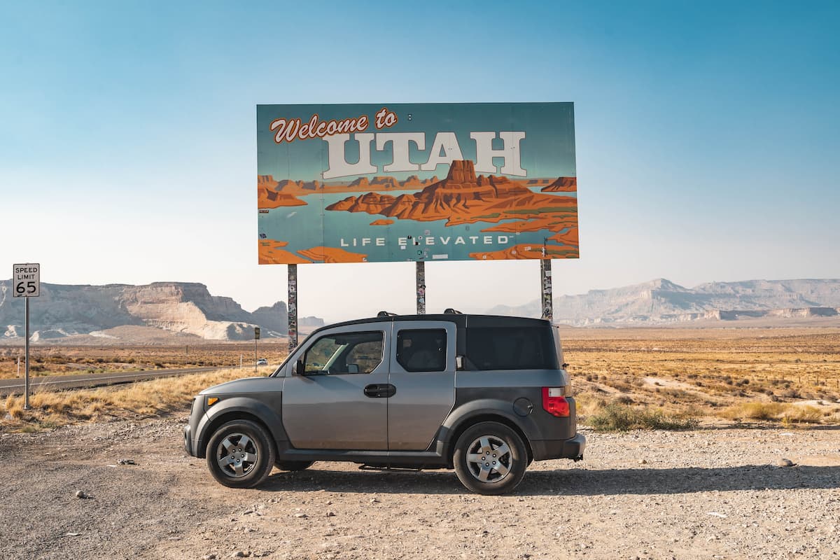 A gray Honda Element parked in front of Welcome to Utah signage.