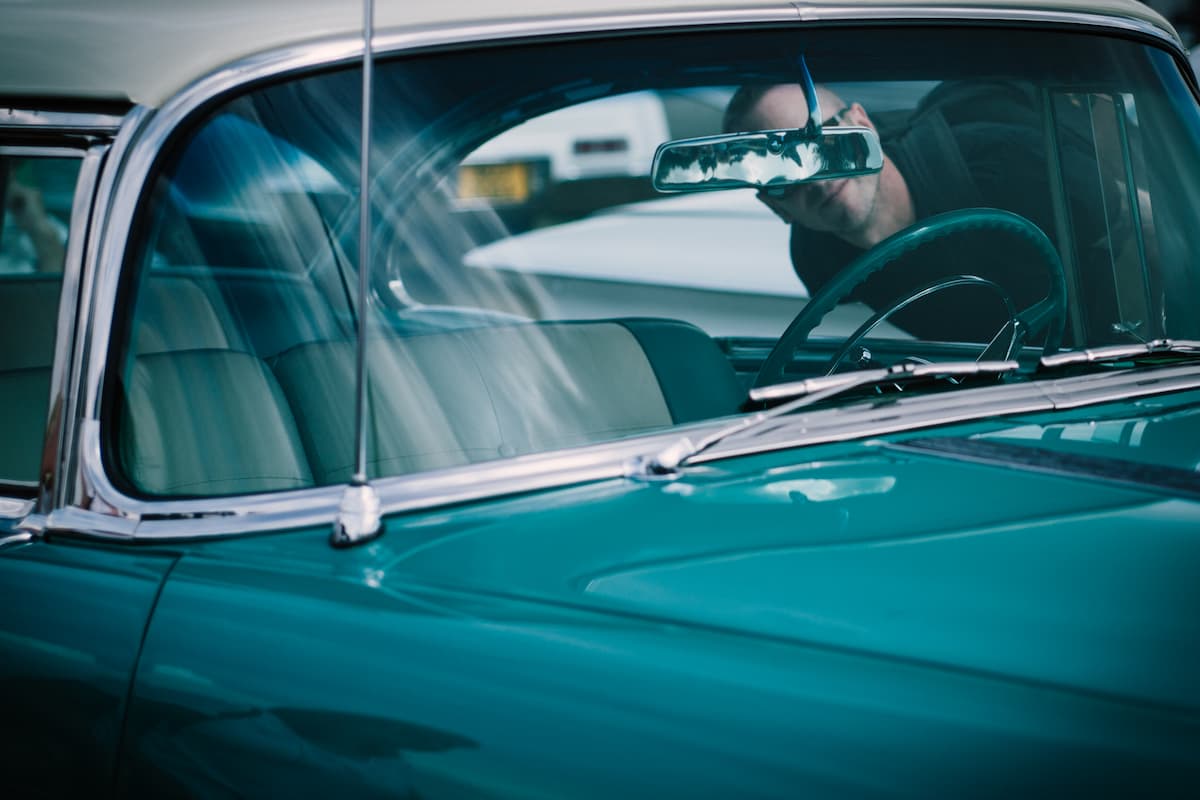 A cropped photo of a blue car with an antenna and a man looking inside the car. 