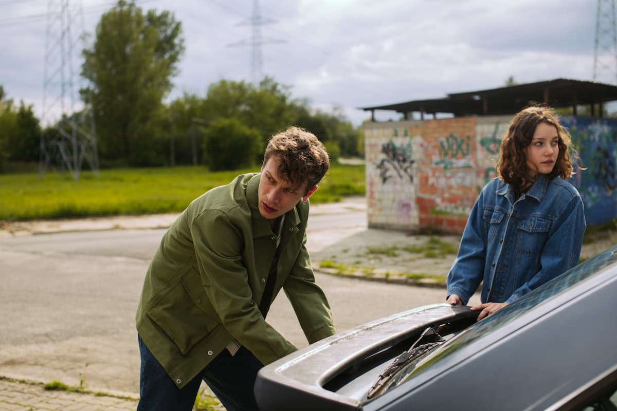 A man in a green jacket and a woman in a denim jacket are looking in different directions while opening someone's car. 