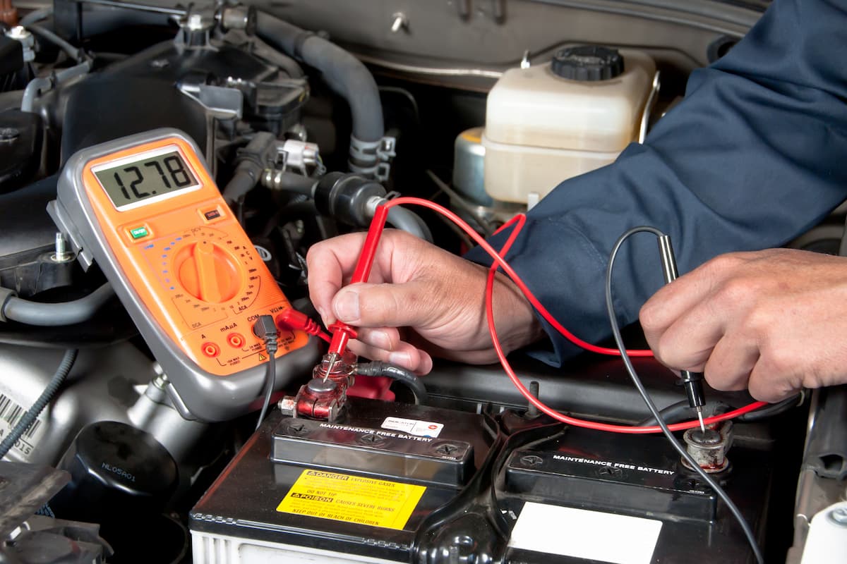 A mechanic checks the car's battery using an orange multimeter.