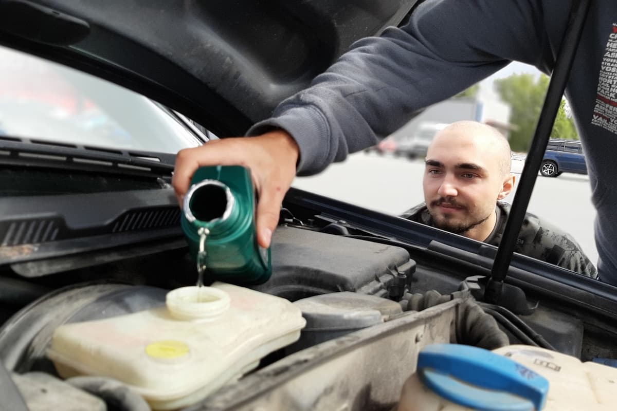 A man looking at the engine oil while another man is pouring it into the car. 