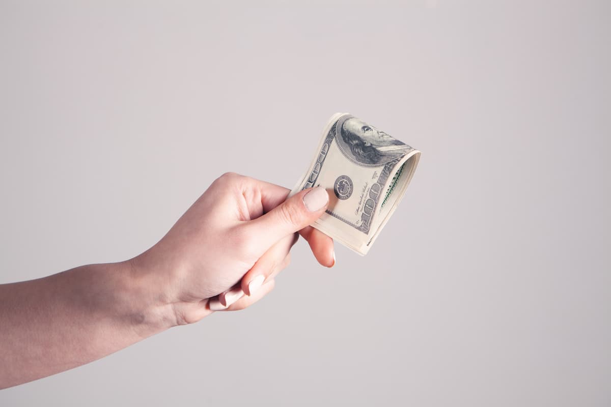 A woman's hand holding a dollar bill near a white wall. 
