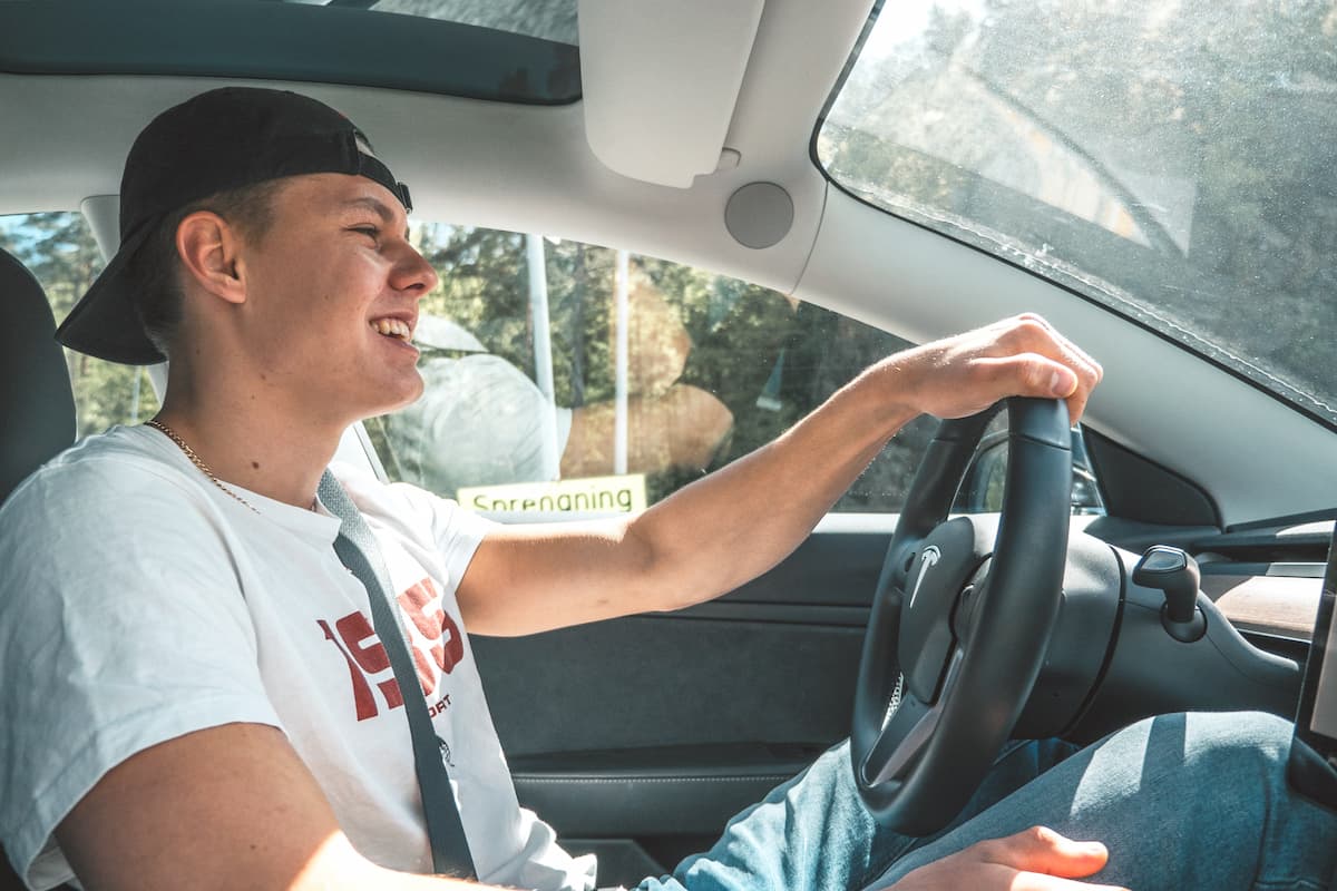 A man in a white t-shirt and black cap is driving a car while smiling. 