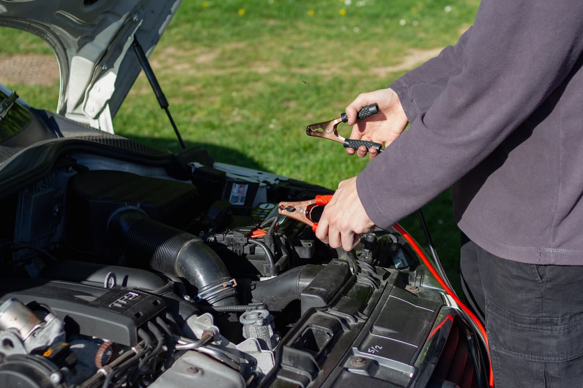 A man's hands clutching jumper cables near a car with its engine visible.