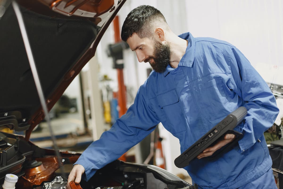 A man in blue uniform is checking the car while holding a diagnostic tool.