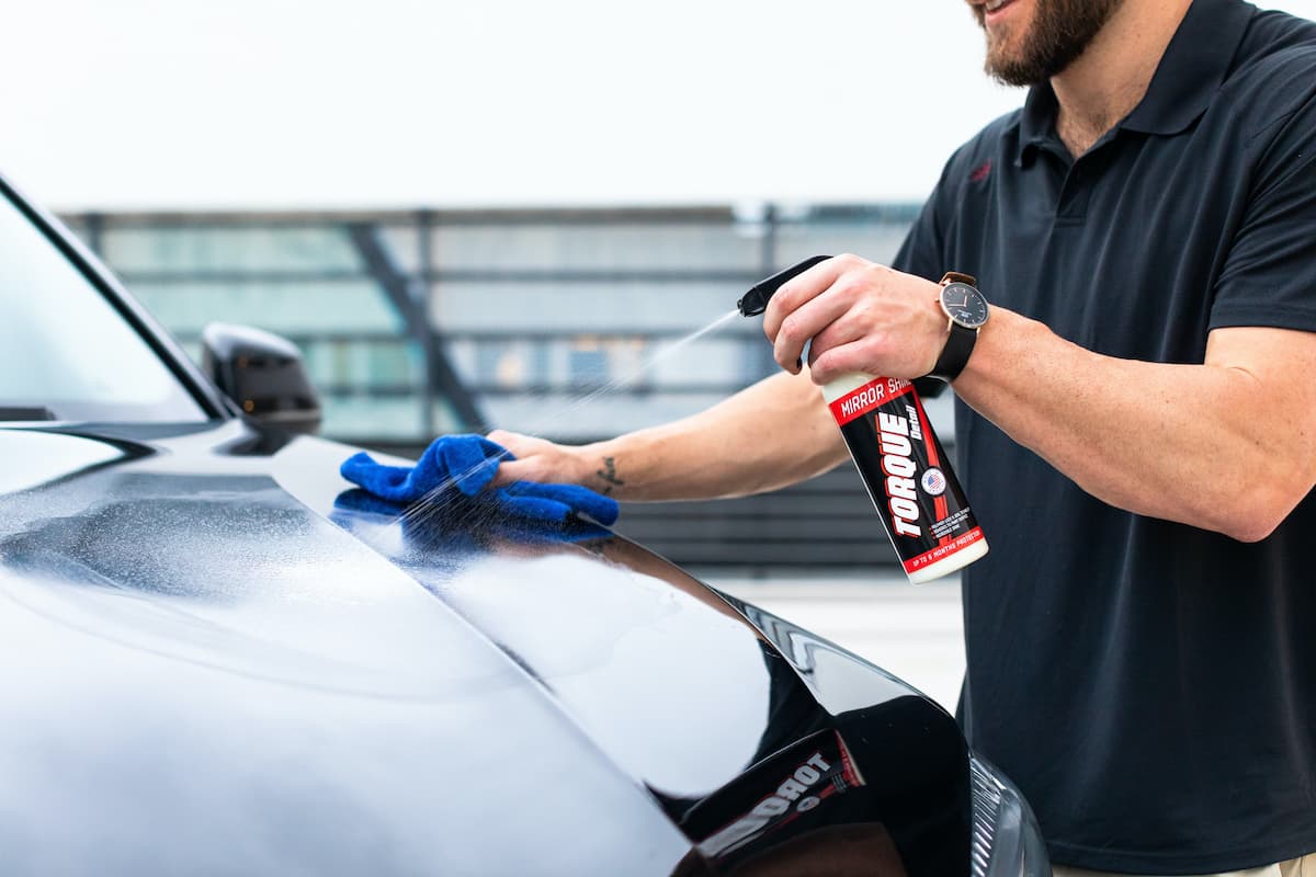 A man in a black polo shirt cleaning the car's hood with Torque Detail Mirror Shine. 