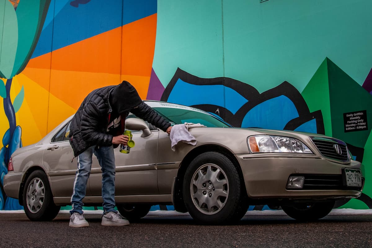 A man in a black jacket and a black hood is wiping a gold car with a towel while holding a green container beside a mural.