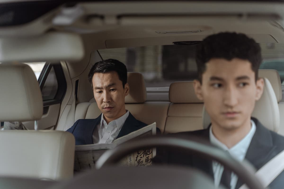 A man in a suit reading a newspaper and a driver sitting in front of the car. 