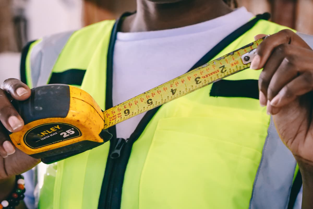 A man wearing a green safety vest is holding a measuring tape.