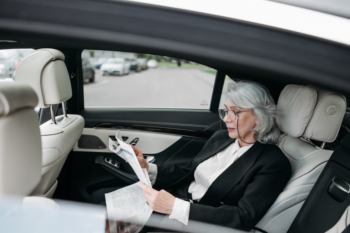 An old woman wearing a black blouse and eyeglasses is reading a newspaper inside a car. 