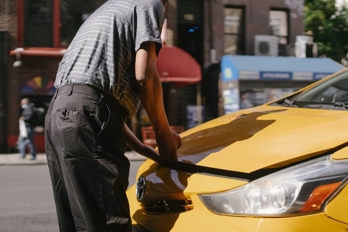 A man opening the hood of a yellow car in the street.