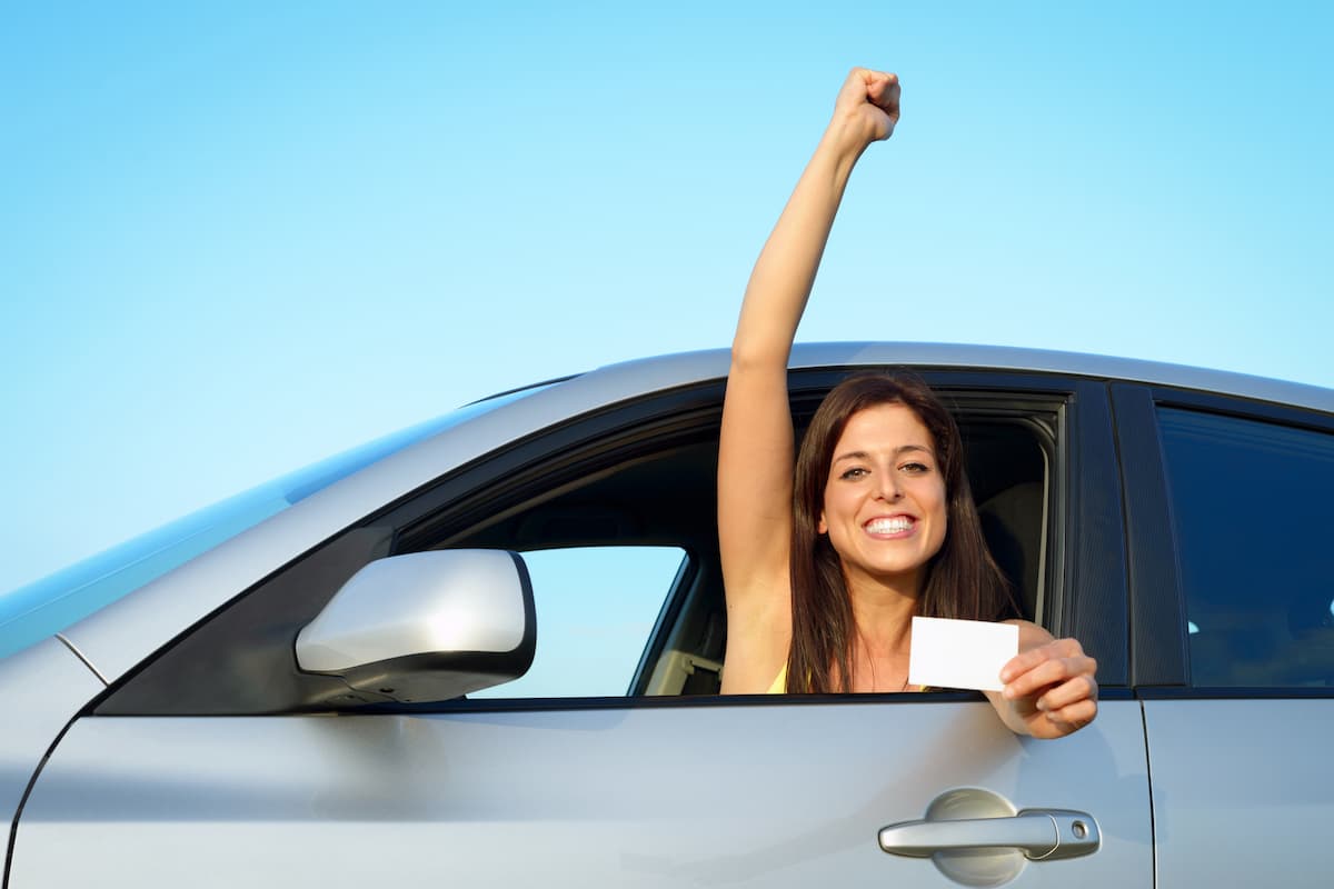 A woman in a car raising her hand and displaying a white blank card after passing her driving test.