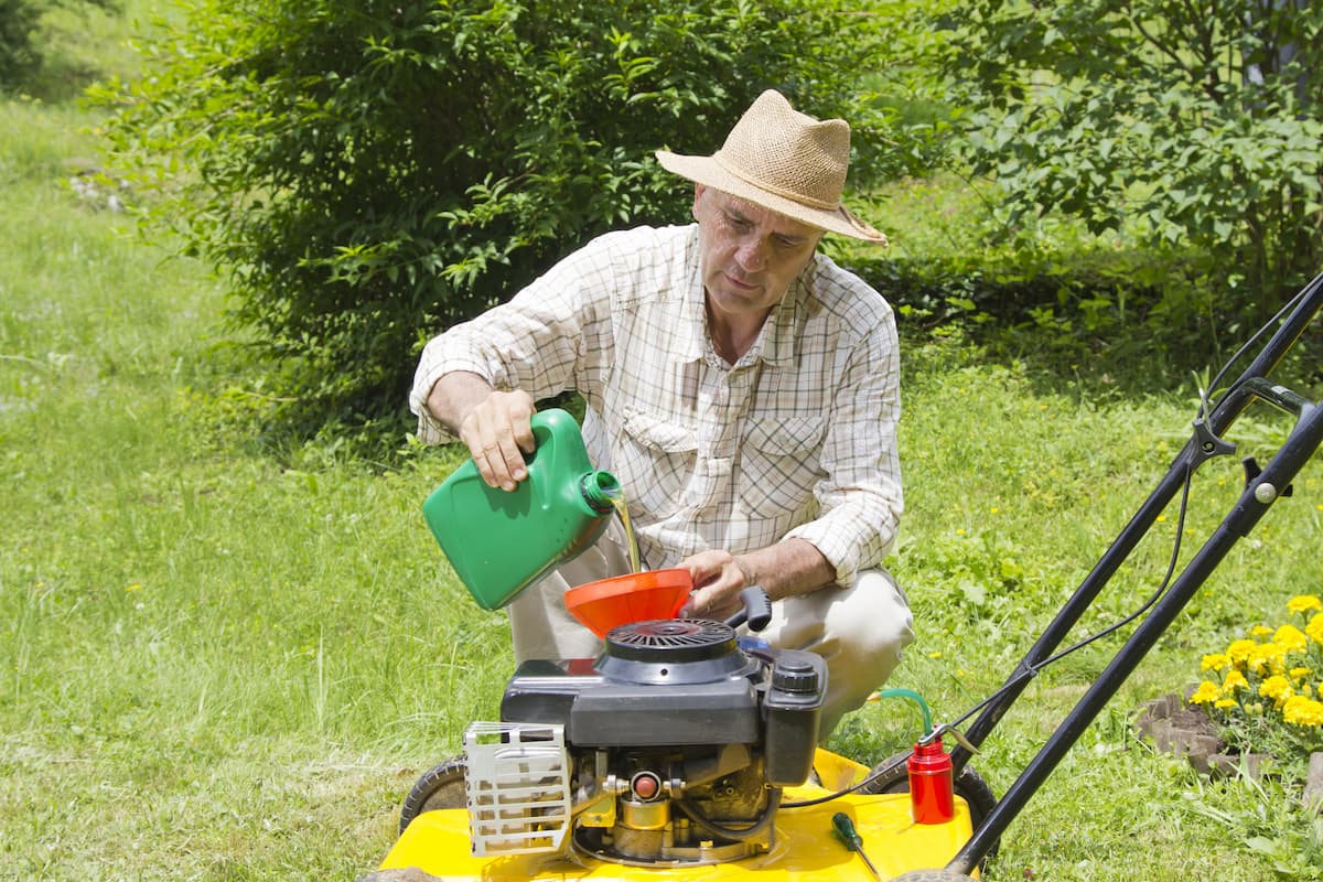 An old man wearing a hat and plaid shirt is putting oil in the lawn mower's engine. 