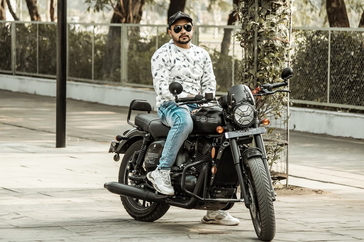 A man in white sweatshirt, black sunglasses and black cap is riding a black motorcycle. 