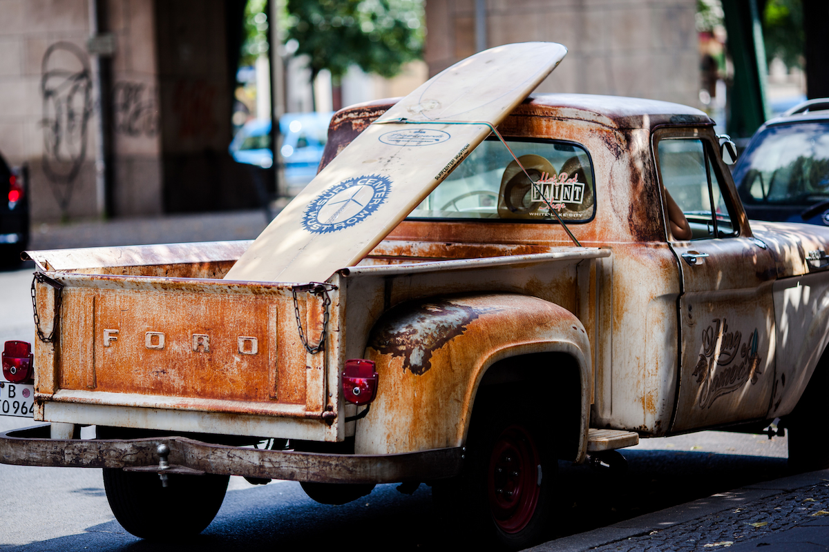 A rusted vintage Ford pickup truck with an old surfboard tied to the truck bed.