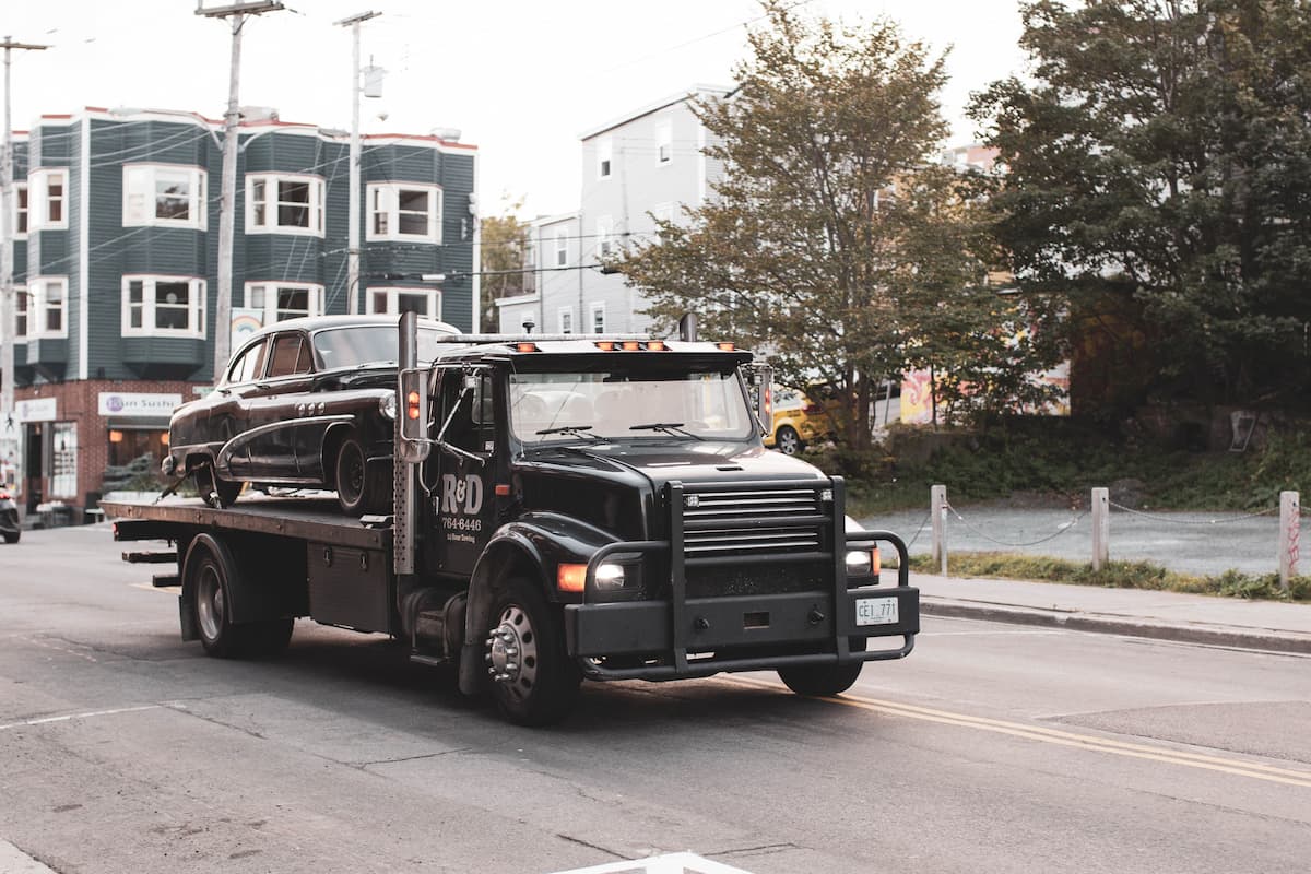A black flatbed tow truck carrying a vintage car while moving on the road. 