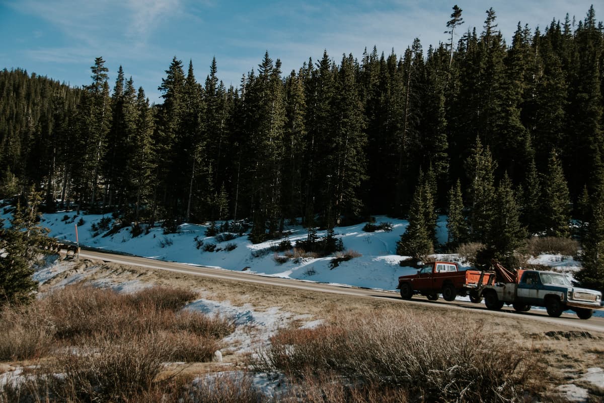 A tow truck towing a pickup truck on a snowy highway surrounded by tall trees.