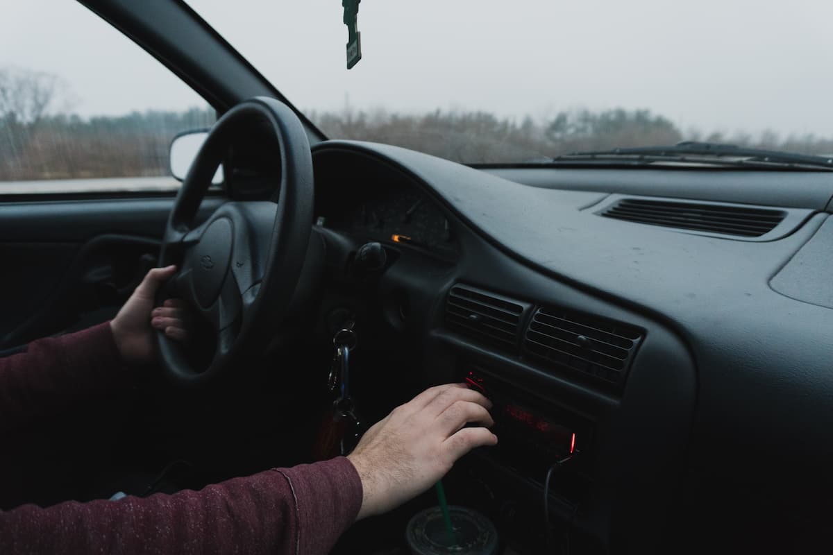 A man holding the steering wheel while turning the car radio. 
