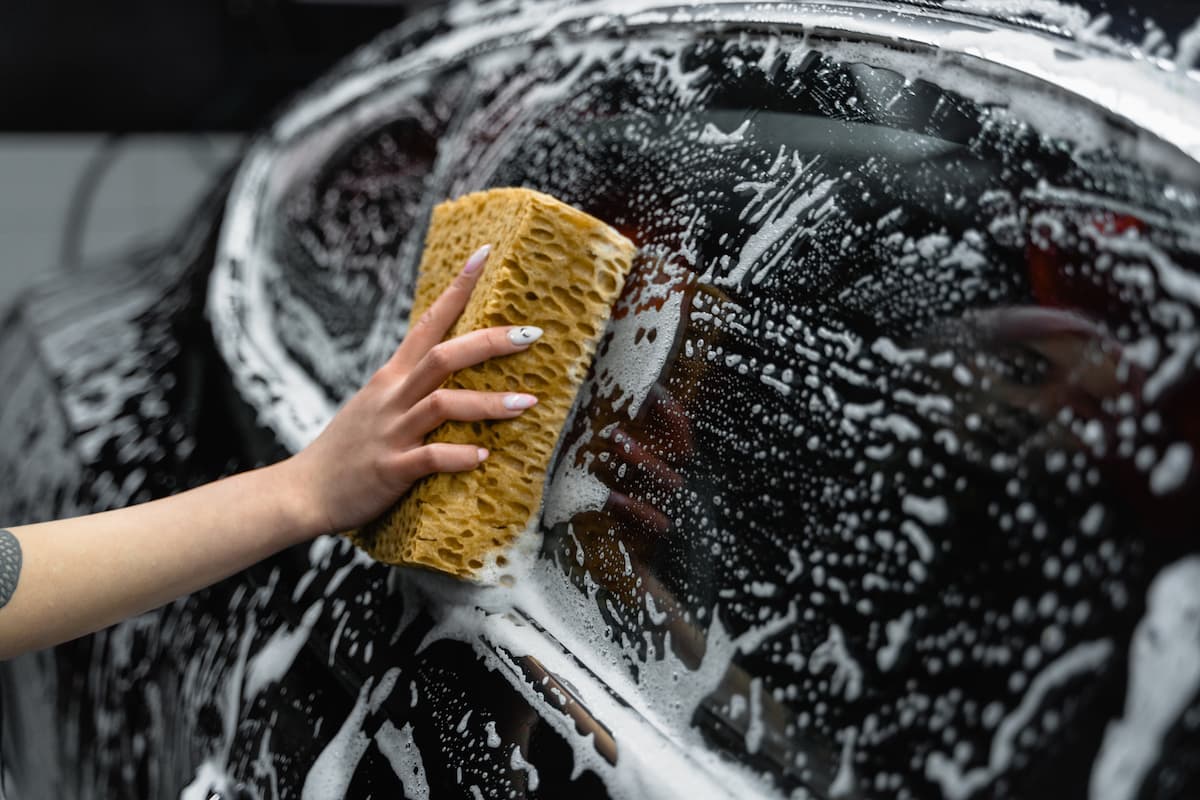 Photo of a woman's hand washing a black car's window with a sponge. 