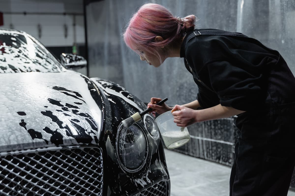 A woman with pink hair is brushing the headlight of a black car. 