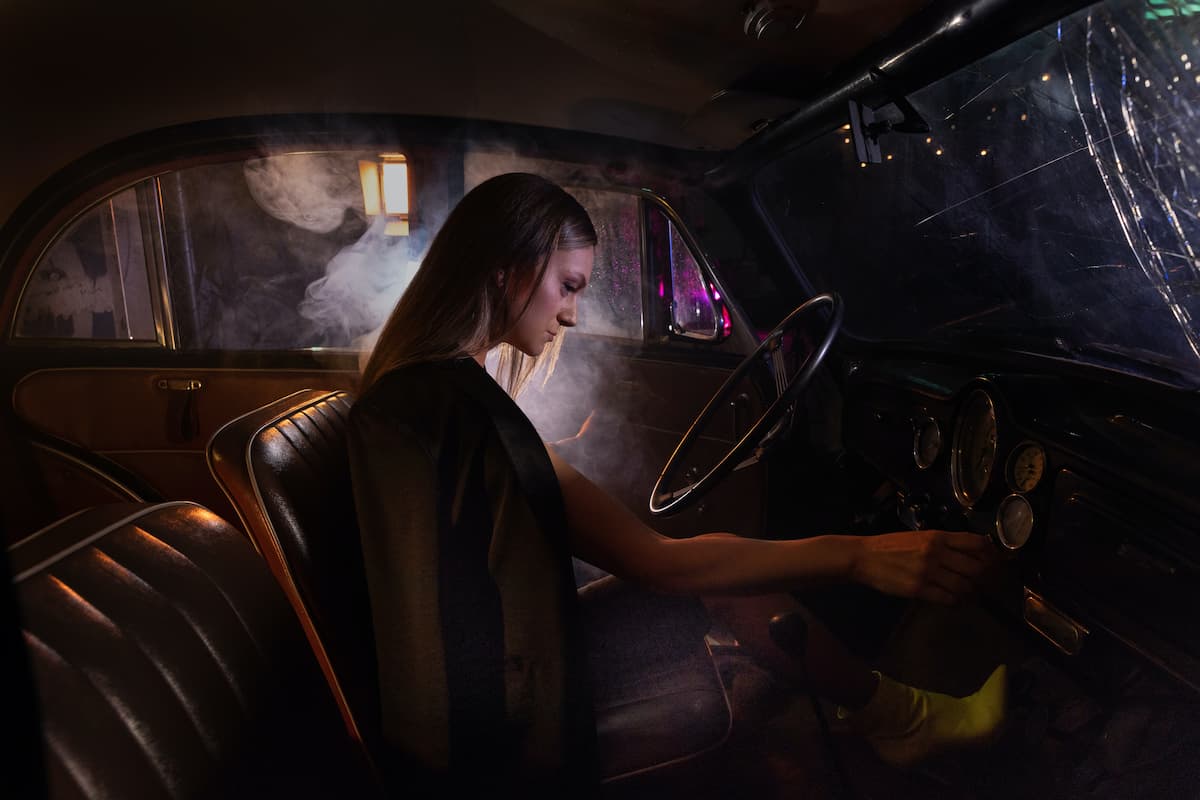 A woman in a coat and yellow boots starts the engine of her car at night.